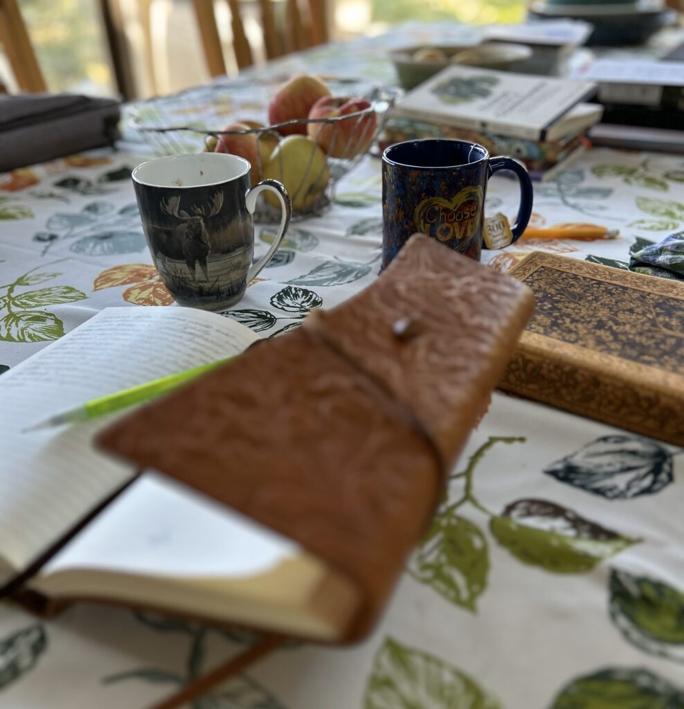 A table with an open journal in the foreground, two cups, a closed journal and books scattered about.